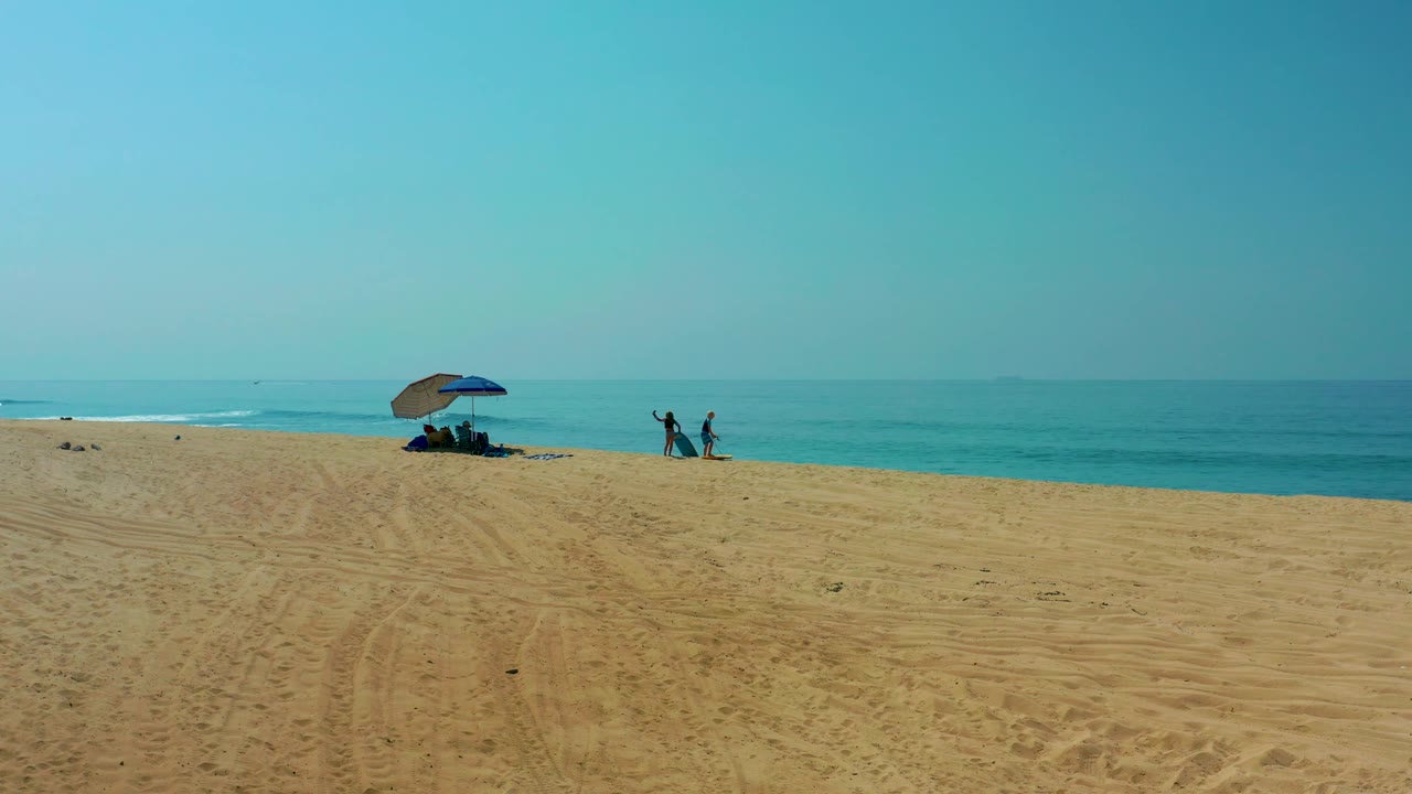At Shipwrecks Los Cabos, two beachgoers stand to the right of the umbrella near the turquoise water, with one or both arms lifted against the open sky.