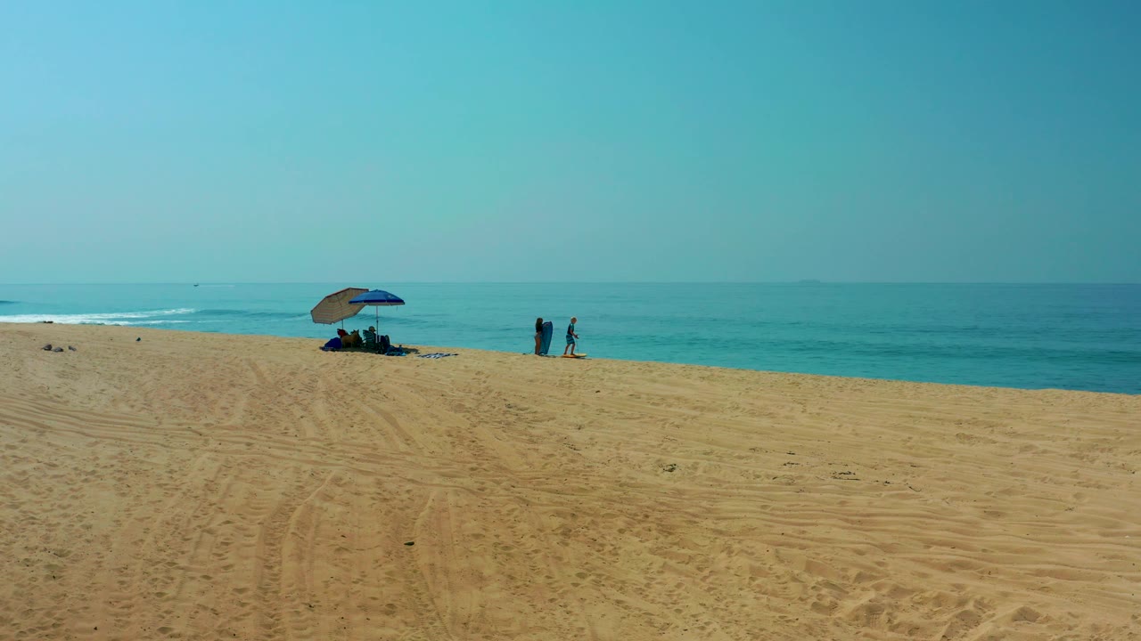 Blue umbrella on sandy beach with two people walking near the ocean.