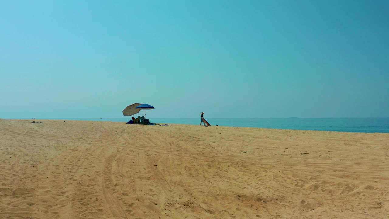 Wide view of Shipwrecks Los Cabos beach with tire-marked sand in front, a blue umbrella shading distant beachgoers, and calm ocean under clear sky.