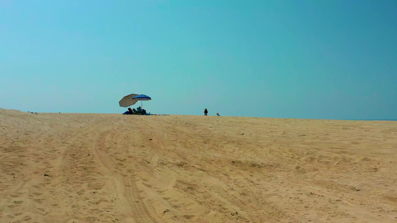 Opening frame at Shipwrecks Los Cabos showing textured sand tracks leading toward a blue umbrella and tiny distant people under a pale blue sky.
