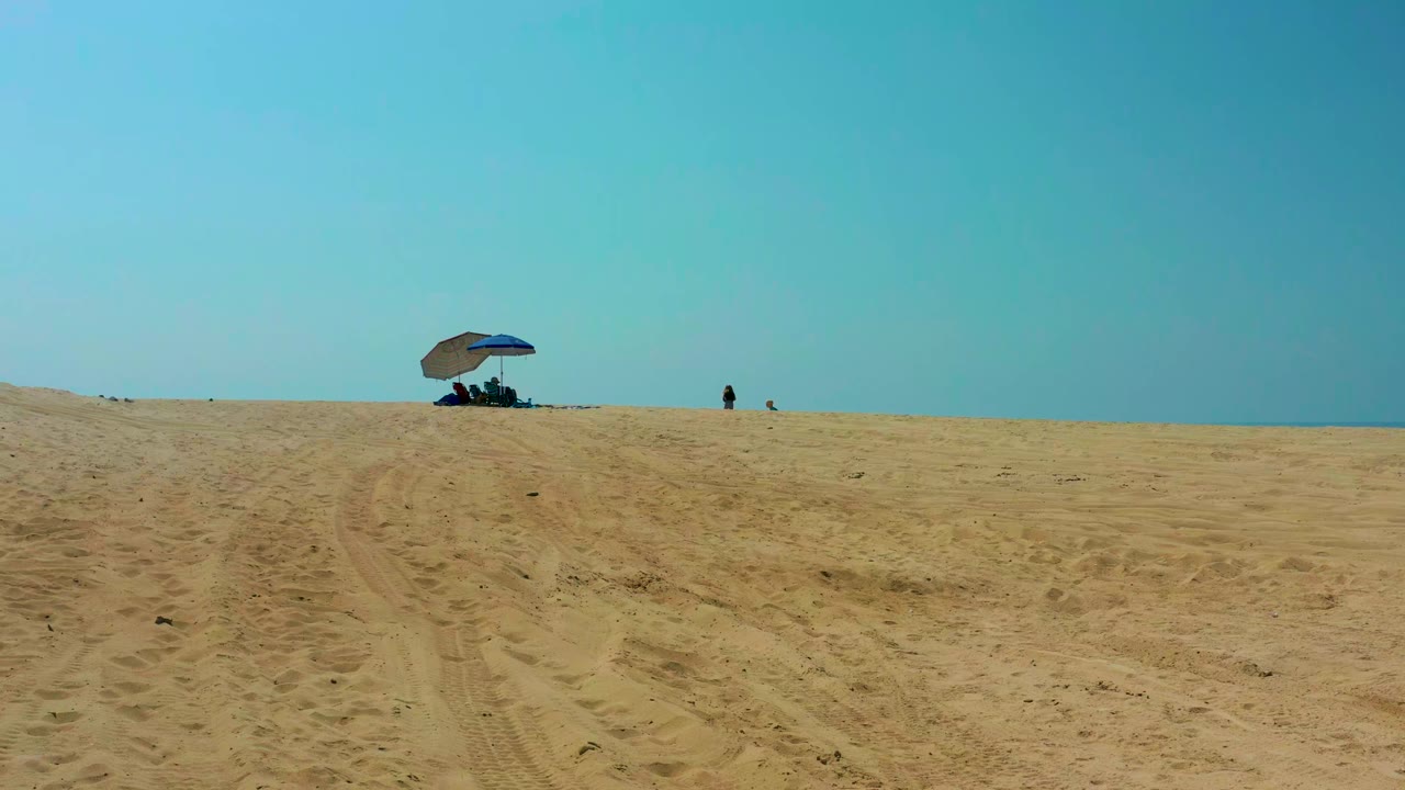 Wide sandy dune with blue umbrella and small figures against a clear sky.