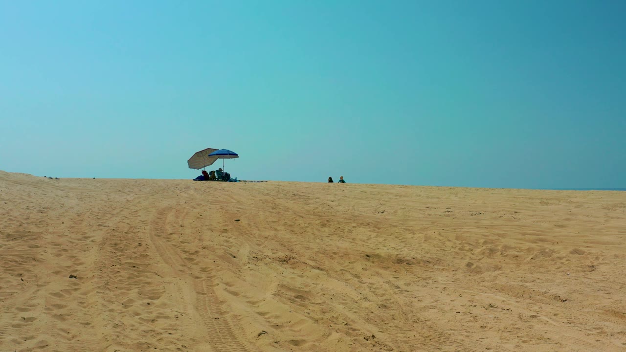 At 0:02, a broad foreground of rutted sand leads to the blue umbrella on the ridge, with tiny beachgoers visible near the horizon in Shipwrecks Los Cabos.