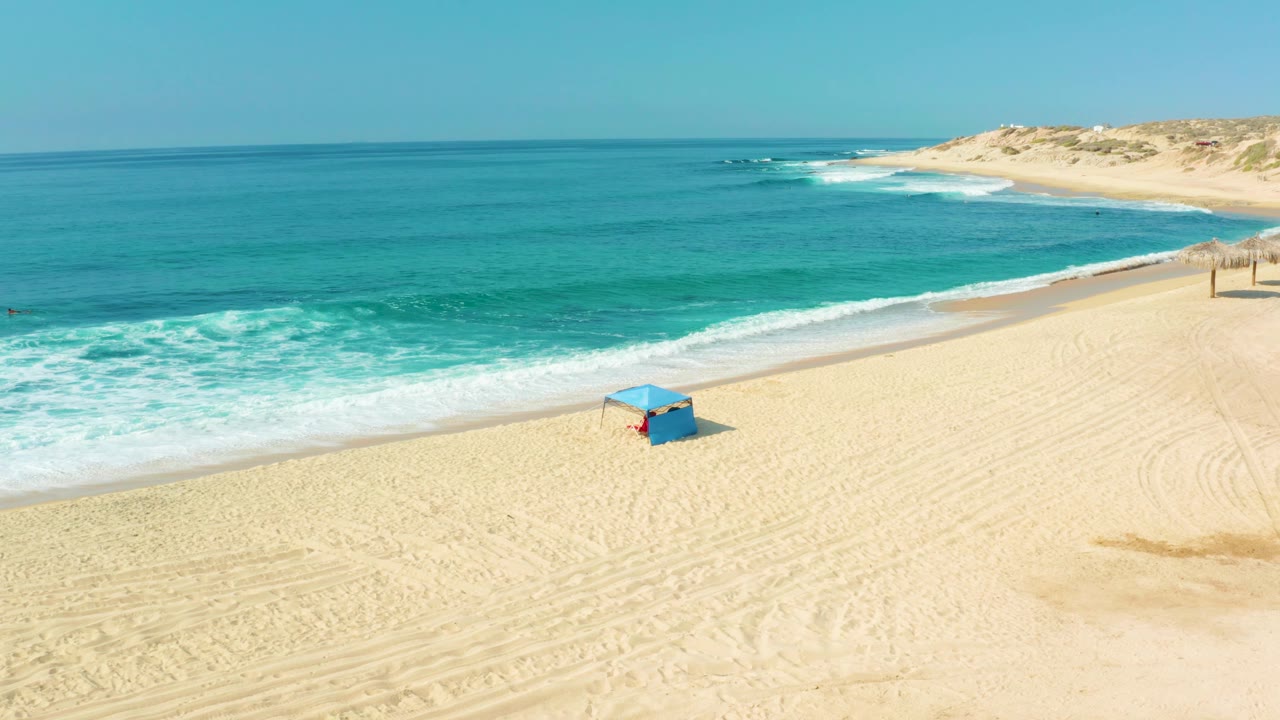 Aerial view at Shipwrecks Los Cabos with the blue hut centered lower in frame, white surf closer to shore, pale tire-marked sand, and no visible people.