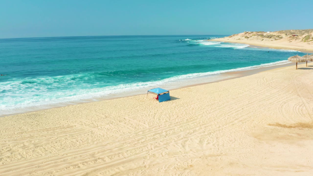 Aerial beach frame from Shipwrecks Los Cabos with the blue hut right of center, turquoise water filling the upper half, low dunes at the far shore, and no visible people.