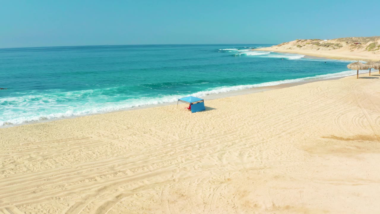 Wide aerial frame at Shipwrecks Los Cabos showing the blue hut lower right of center, foamy waves entering from the left, empty sand with tire marks, and no visible people.
