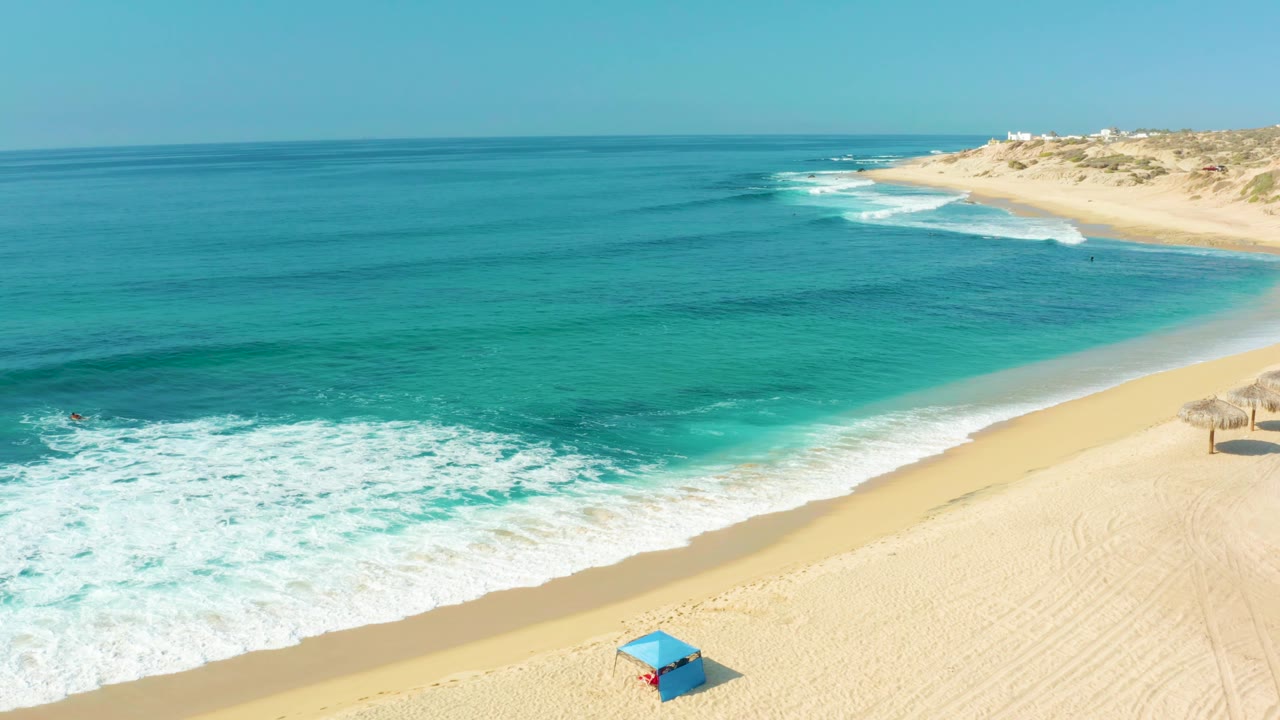 Aerial frame at Shipwrecks Los Cabos with a broad band of white foam pushing toward the sand, open turquoise water offshore, distant dunes, and no visible people.