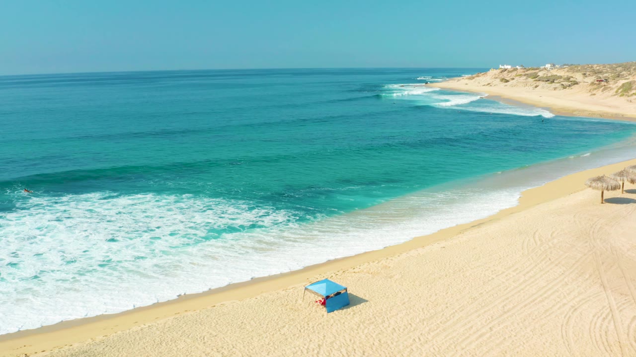 Aerial frame at Shipwrecks Los Cabos showing a long diagonal shoreline, foamy wave wash, empty-looking sand, and dune-backed coast under a clear sky, with no visible people.