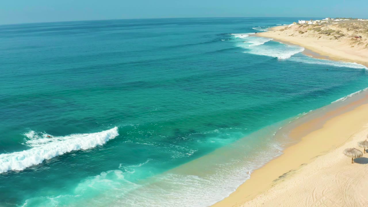 Aerial frame at Shipwrecks Los Cabos with white surf advancing across turquoise water toward pale sand, distant dunes, and no visible people.