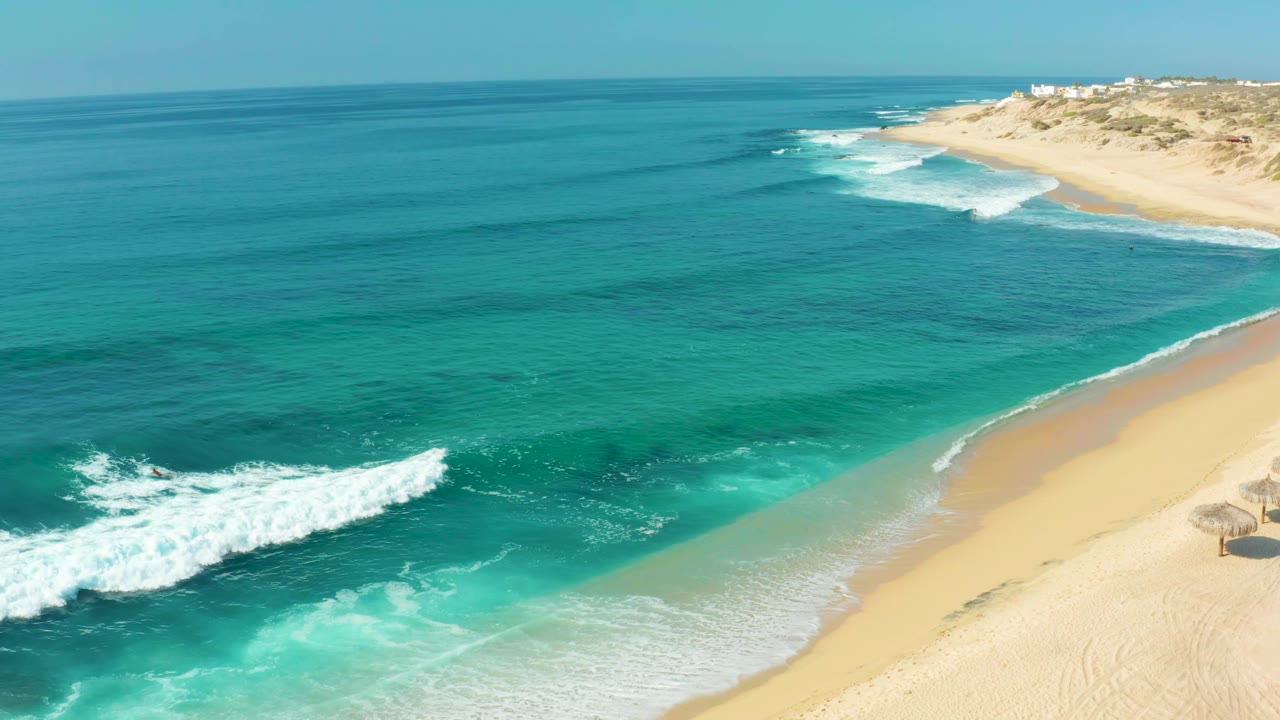 Aerial view of Shipwrecks Los Cabos with a larger white wave curling into the turquoise shallows beside the curved sandy shoreline, with no visible people.