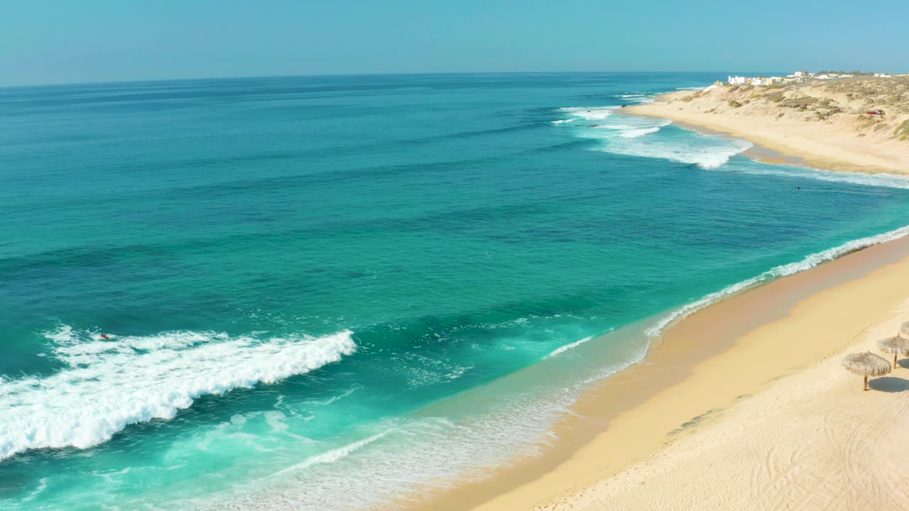 Wide aerial frame at Shipwrecks Los Cabos showing open blue water, turquoise nearshore surf, pale sand, and distant beach dunes, with no visible people.