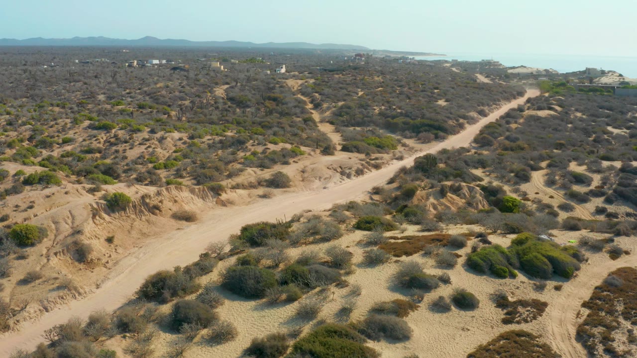 Aerial view over Shipwrecks Los Cabos showing a sandy dirt road curving through low desert shrubs, distant mountains, a pale sky, coastline on the horizon, and no visible people.