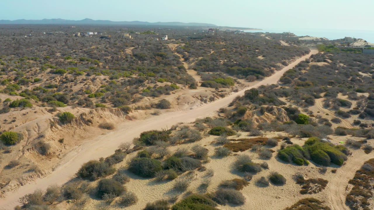 Elevated view at Shipwrecks Los Cabos showing a pale dirt road cutting through dry scrub, sand mounds in the foreground, a small vehicle on the track, mountains in the distance, coastal water near the horizon, and no visible people.