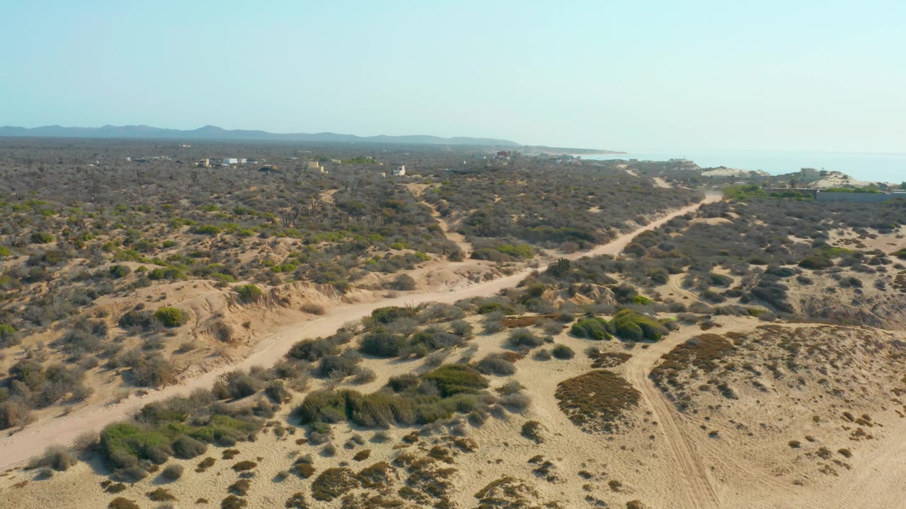 Aerial view in Shipwrecks Los Cabos with a pale dirt road cutting through scrub-covered sandy terrain, ocean visible to the right, distant mountains, and no visible people.
