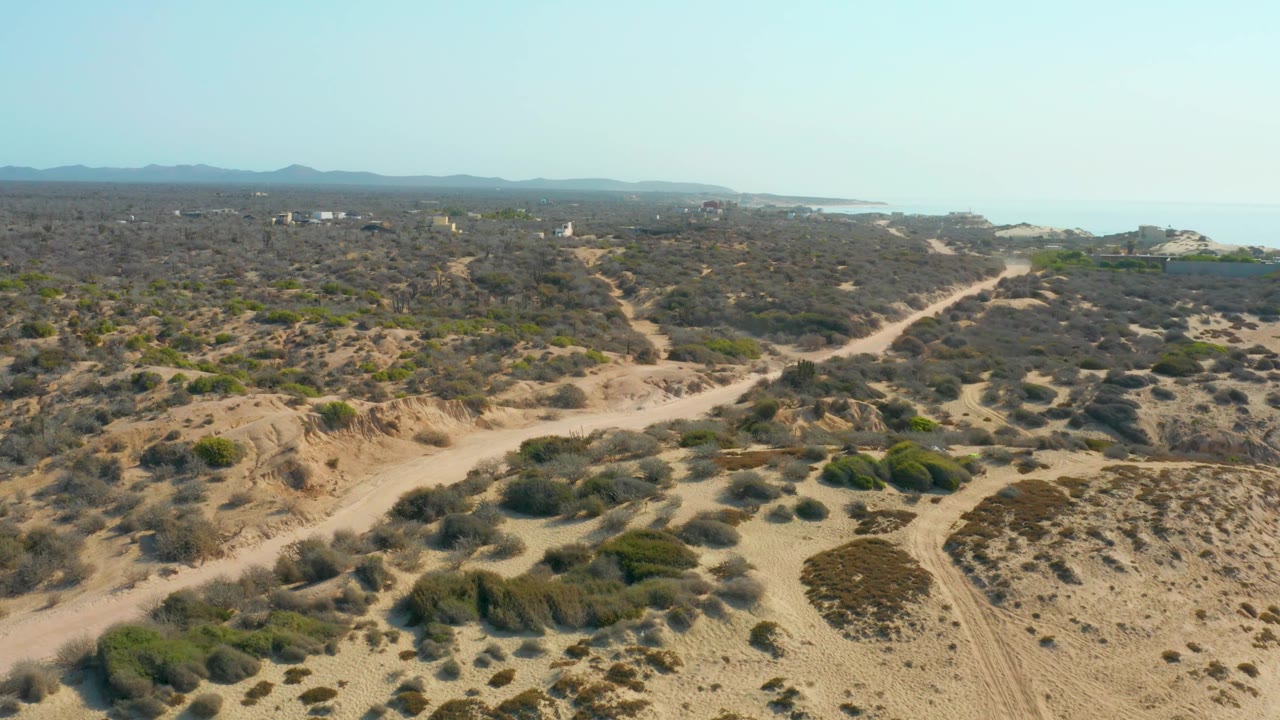 Wide frame over Shipwrecks Los Cabos showing dry sandy hills with scattered brush, intersecting dirt tracks, the ocean band on the right horizon, mountains beyond, and no visible people.