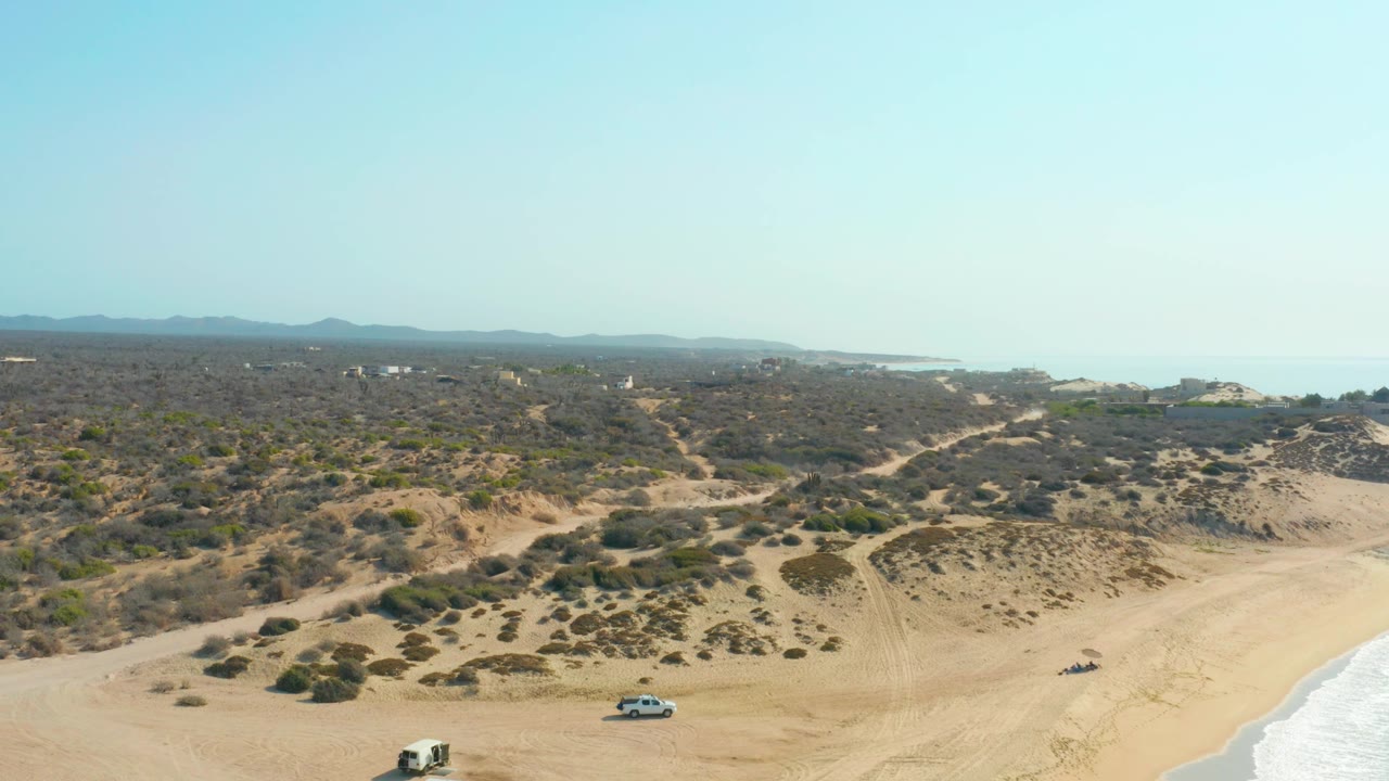 Aerial view at Shipwrecks Los Cabos with the white SUV slightly farther right along the lower beach, the dark vehicle near the shoreline side, scrub dunes, ocean in the background, and no visible people.