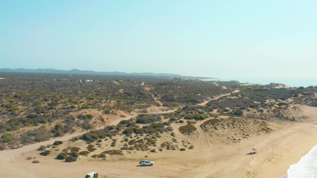 Frame from Shipwrecks Los Cabos showing a white SUV near the lower center-left beach, a smaller dark vehicle on the right sand, winding dune tracks, low vegetation, ocean, and no visible people.