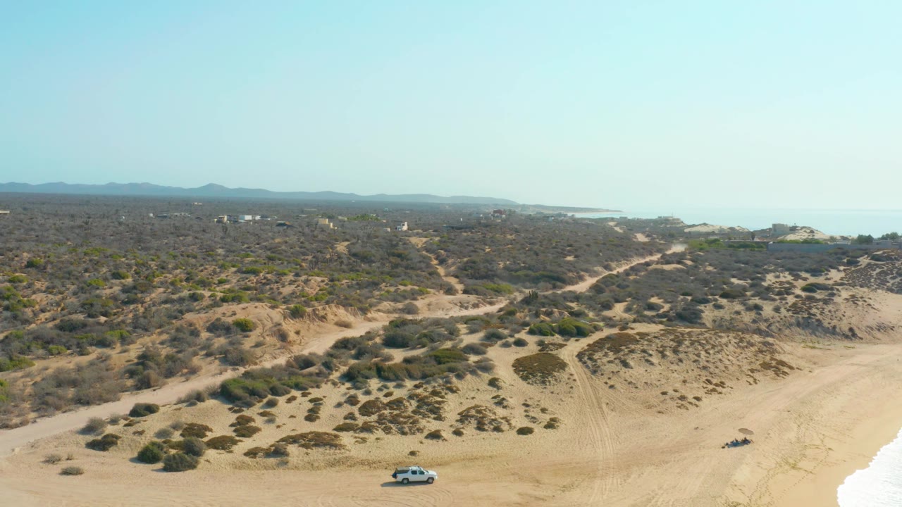 Shipwrecks Los Cabos aerial frame with the white SUV lower left of center, a dark vehicle on the right beach, pale sand foreground, scrubby dune ridges, distant water, and no visible people.