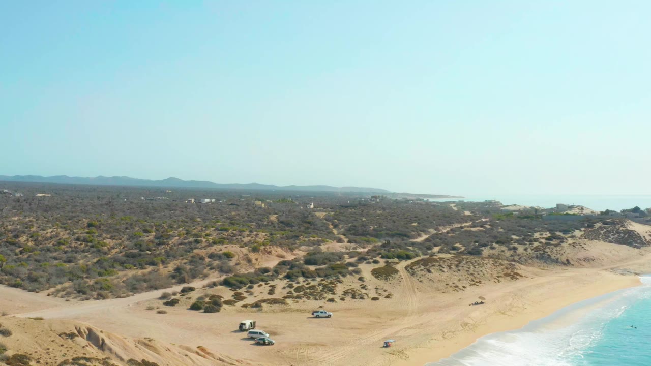 Shipwrecks Los Cabos beach seen from above with the vehicle cluster slightly left of center, a single vehicle farther along the shore, scrubby dunes, and no visible people.