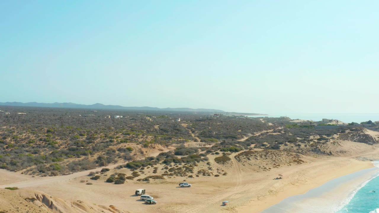 Aerial frame at Shipwrecks Los Cabos with vehicles lower in the shot, dune ridges and brush across the middle, a pale horizon, ocean at the right, and no visible people.