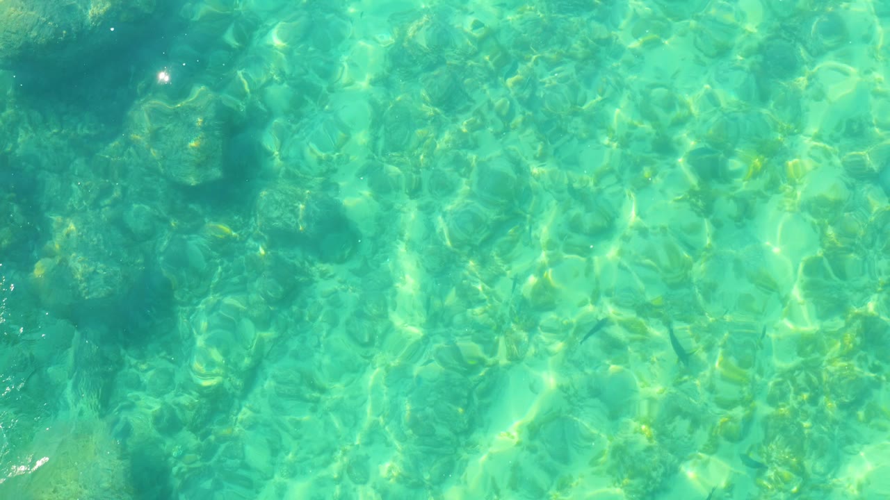 Overhead view of clear turquoise water at Shipwrecks Los Cabos with bright ripples over darker submerged rock patches and no visible people.