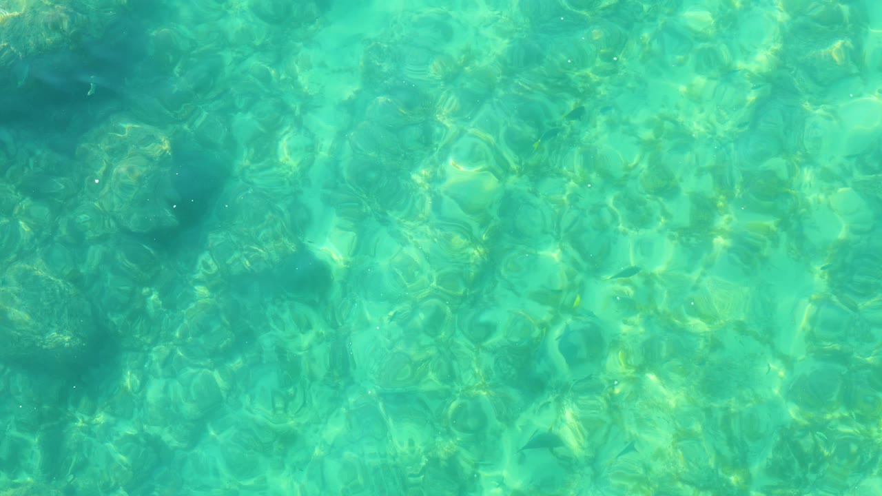 At Shipwrecks Los Cabos, clear turquoise water fills the frame over a shallow rocky bottom, with scattered darker patches beneath the surface and no visible people.