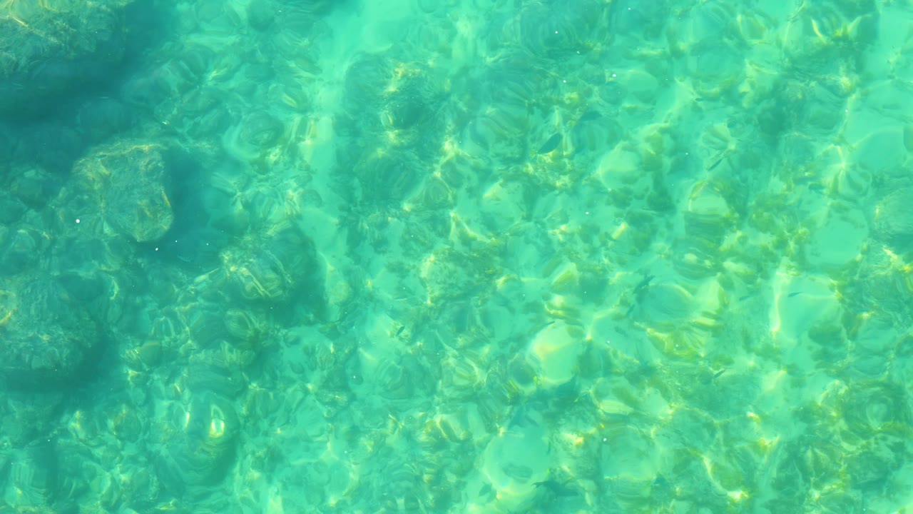 At 0:02, transparent turquoise water at Shipwrecks Los Cabos reveals a darker submerged patch near the upper area and textured seabed below, with no visible people.