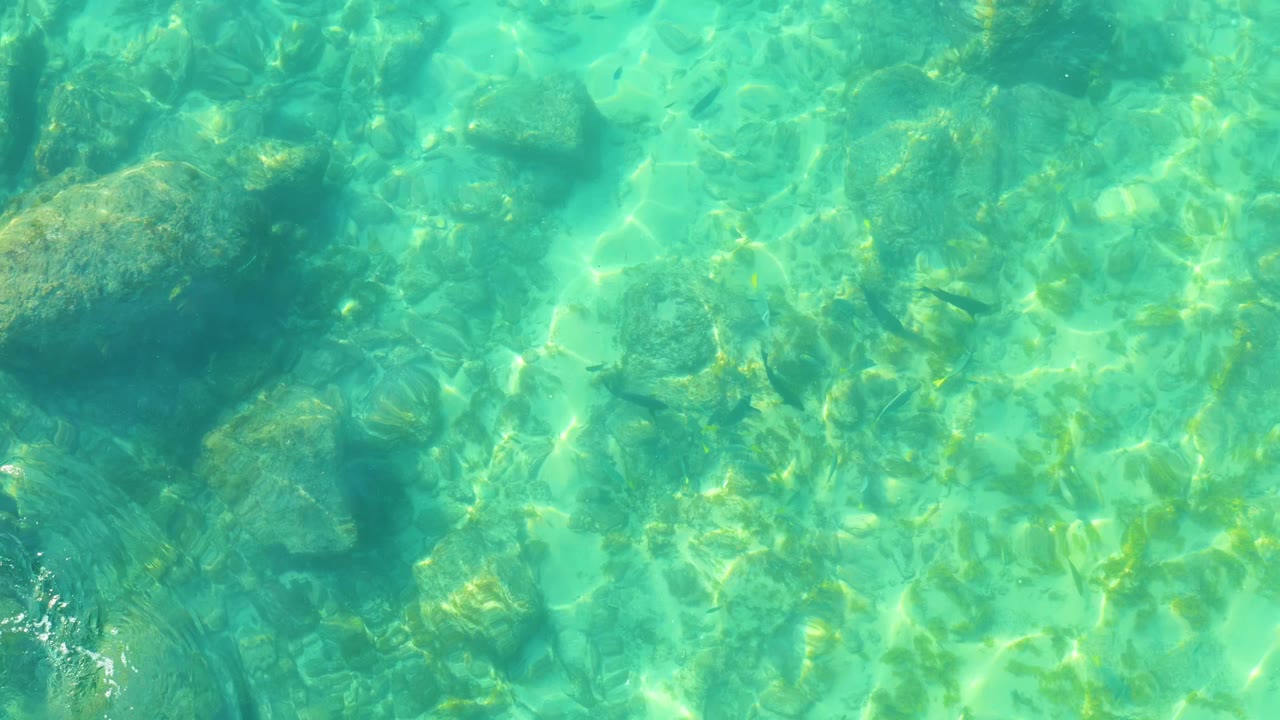 Sunlit shallow water at Shipwrecks Los Cabos shows a large darker rock shape near the upper right and no visible people.