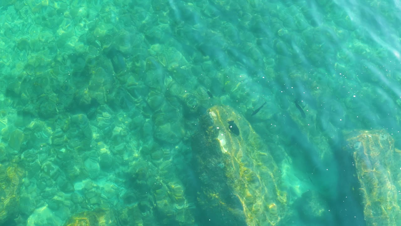 At 0:01, the blue-green water shifts over the rocky bottom, with a large pale rock lower left and no visible people.