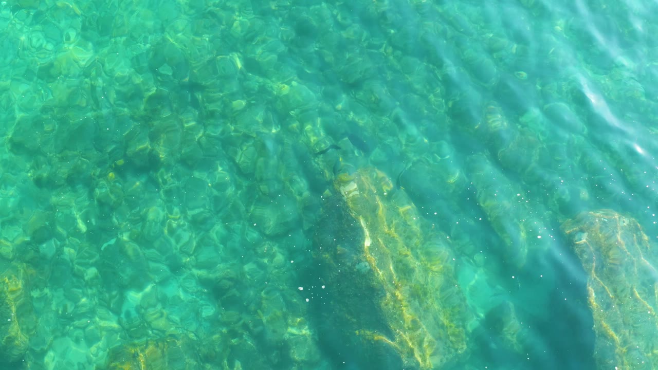 Shallow water over a rocky seabed at Shipwrecks Los Cabos fills the frame, with brighter ripple lines crossing diagonally and no visible people.