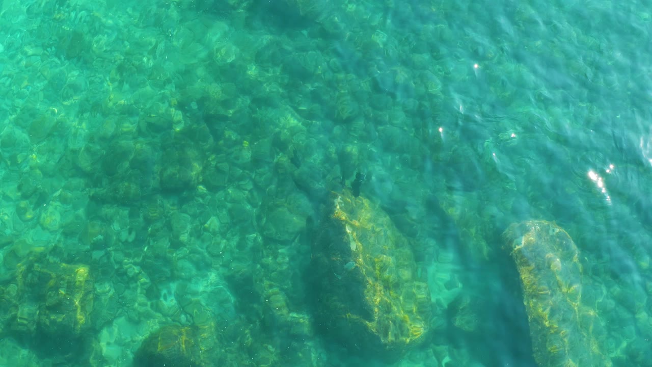 Overhead view of clear turquoise water at Shipwrecks Los Cabos, Baja California Sur, with submerged rocks, green patches, surface ripples, and no visible people.