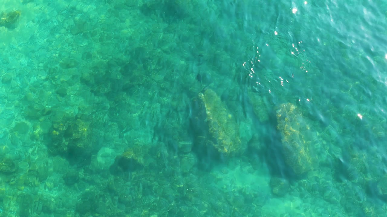 A rocky ocean floor is visible through turquoise rippled water at Shipwrecks Los Cabos, with scattered darker rock patches and no visible people.