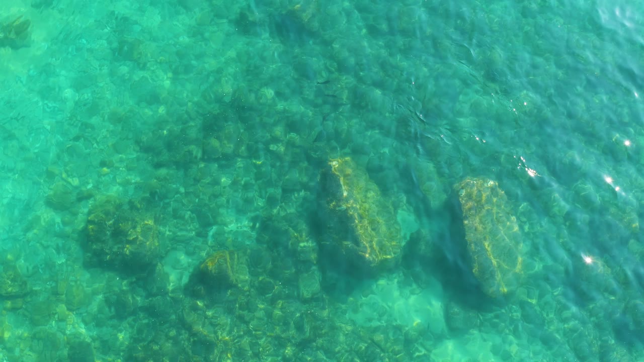 Shallow turquoise water reveals several larger rocks on the seabed at Shipwrecks Los Cabos, with soft wave distortion across the frame and no visible people.