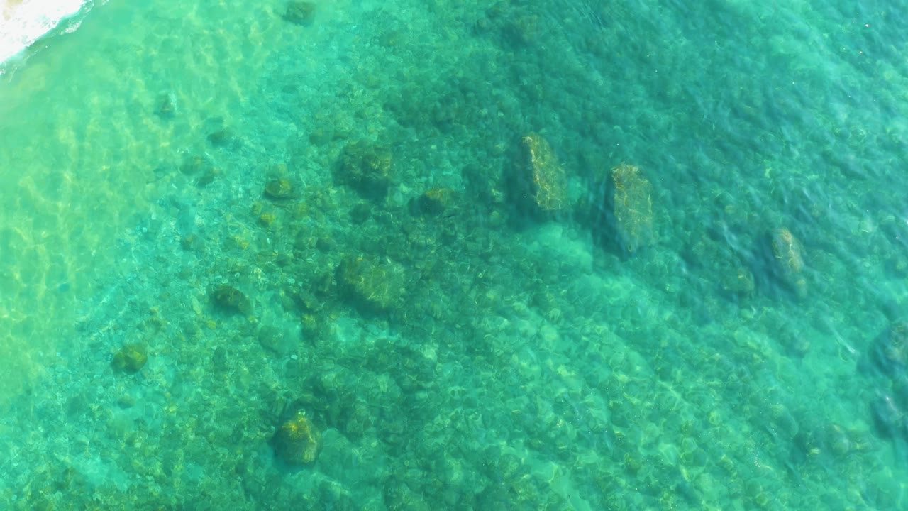 Clear shallow water at Shipwrecks Los Cabos reveals uneven rocks and green marine growth beneath a lightly rippled turquoise surface, no visible people.