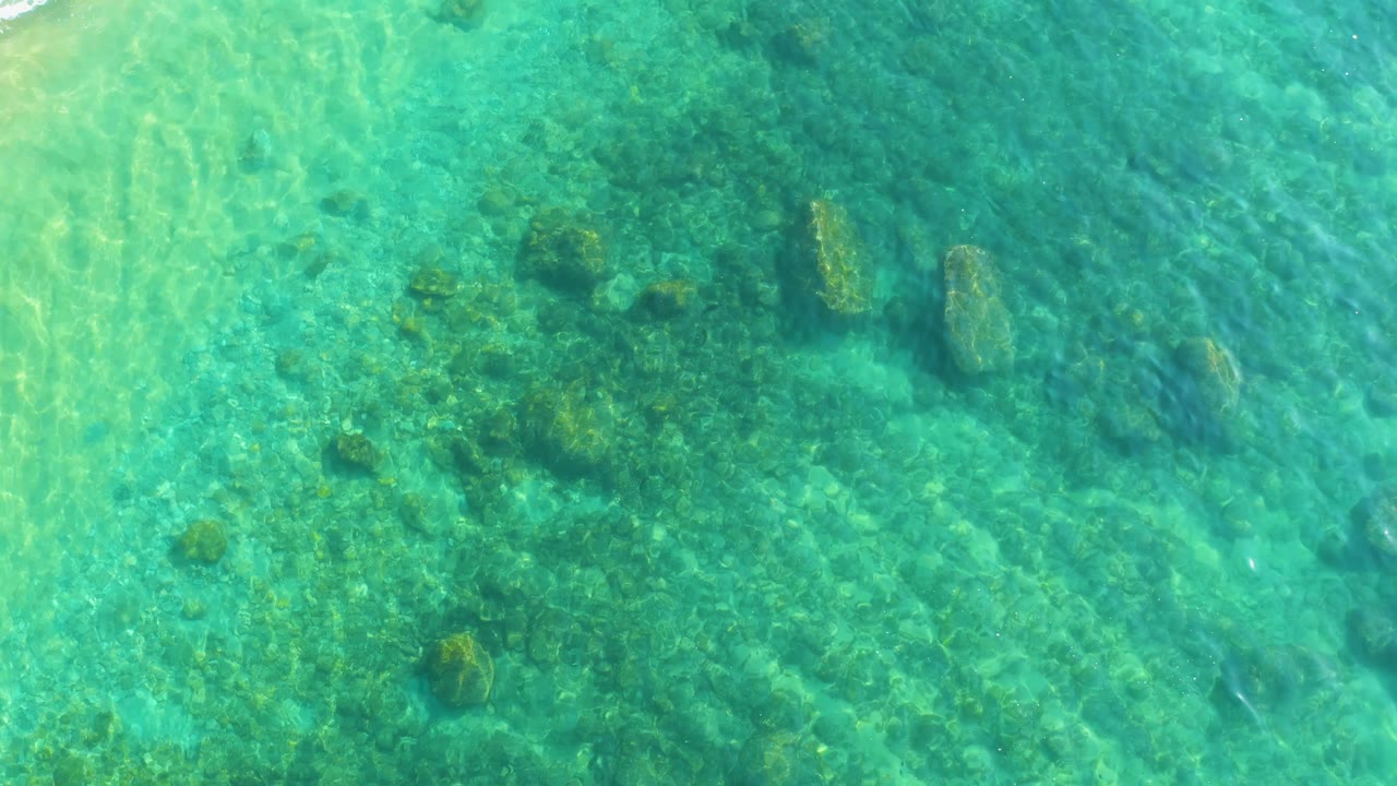 A cluster of submerged rocks sits near the center of the overhead frame at Shipwrecks Los Cabos, with turquoise water and faint surface shimmer around it, no visible people.