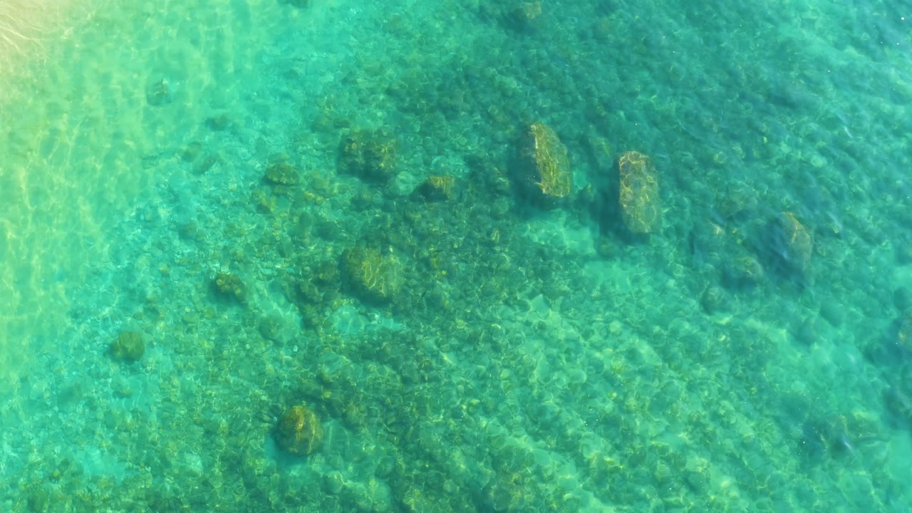 Sunlit turquoise water at Shipwrecks Los Cabos passes over a rocky seabed with green patches and soft wave lines across the frame, no visible people.