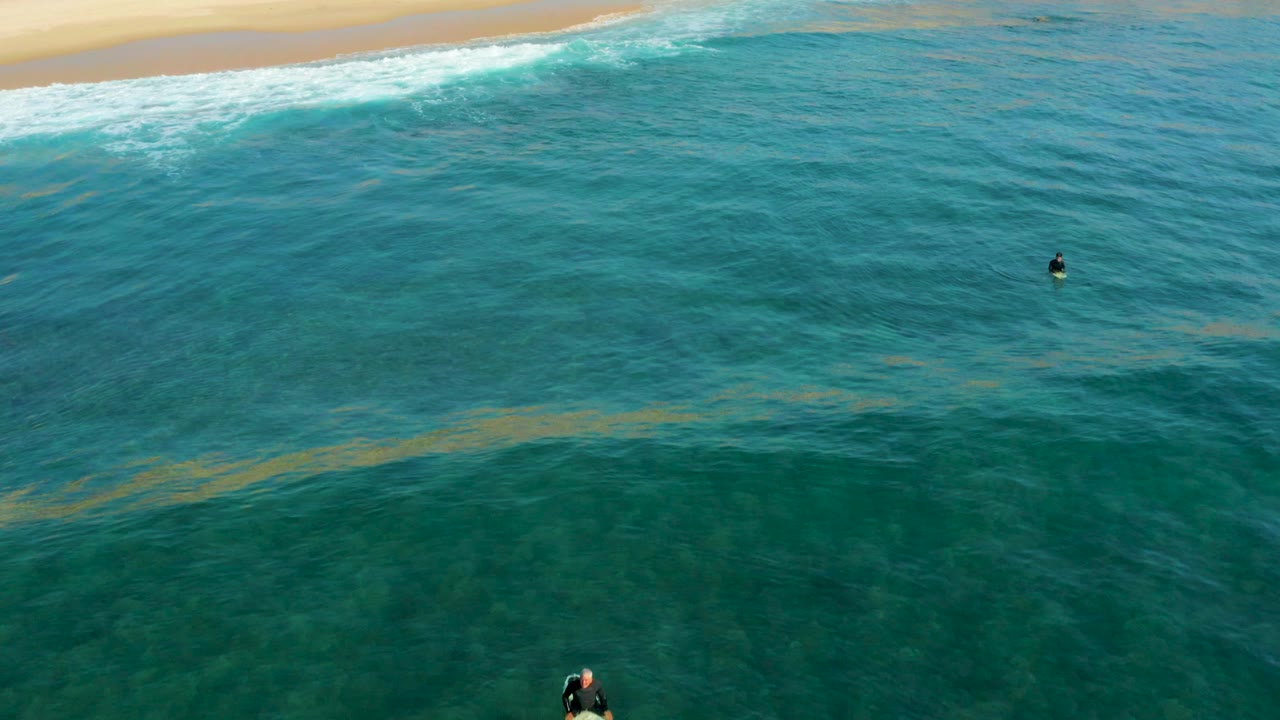 Aerial frame at Shipwrecks Los Cabos showing turquoise water, a sandy shoreline at the top, white surf, and two small swimmers separated across the ocean surface.