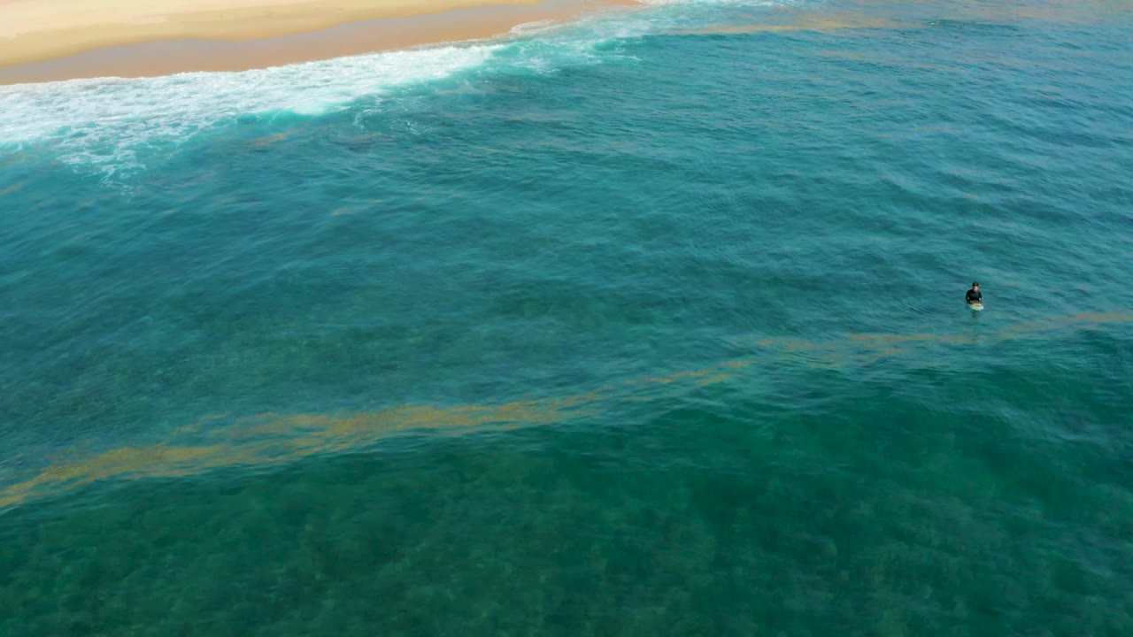 Aerial frame at Shipwrecks Los Cabos showing clear turquoise water, a diagonal surf line near the beach, and one small swimmer near the right edge of the frame.