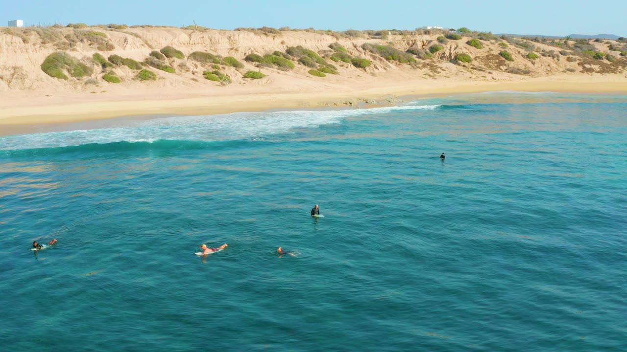 Aerial view at Shipwrecks Los Cabos showing several people spread across clear blue-green ocean water, with a sandy beach and low dunes across the top of frame.