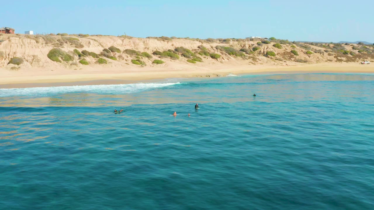 Opening frame of Shipwrecks Los Cabos with rippled blue-green water in the foreground, several tiny swimmers offshore, a sandy beach, and pale cliffs beyond.