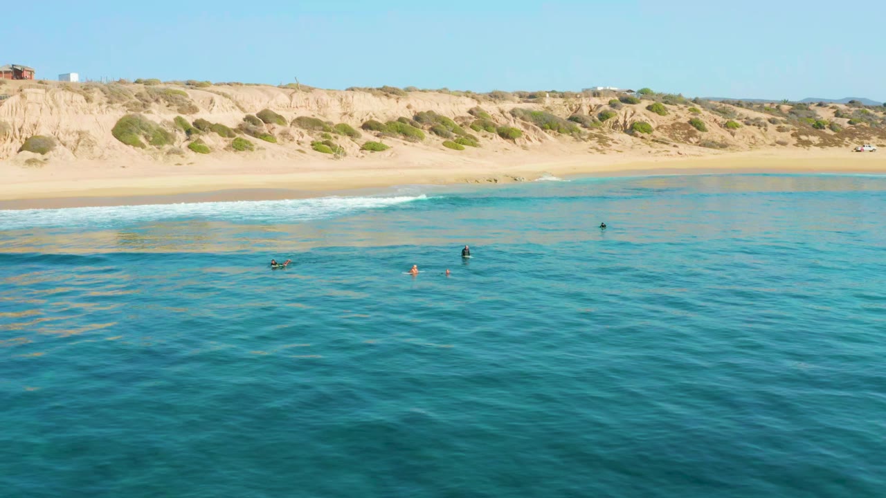 Swimmers in clear ocean water below a sandy beach and cliff line.