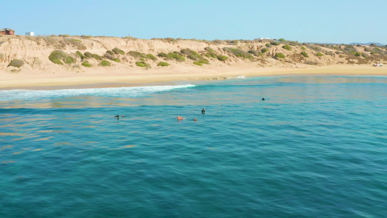 At 0:02, small swimmers appear across the middle of the clear water while a white wave line runs along the pale sand beneath the cliffs at Shipwrecks Los Cabos.