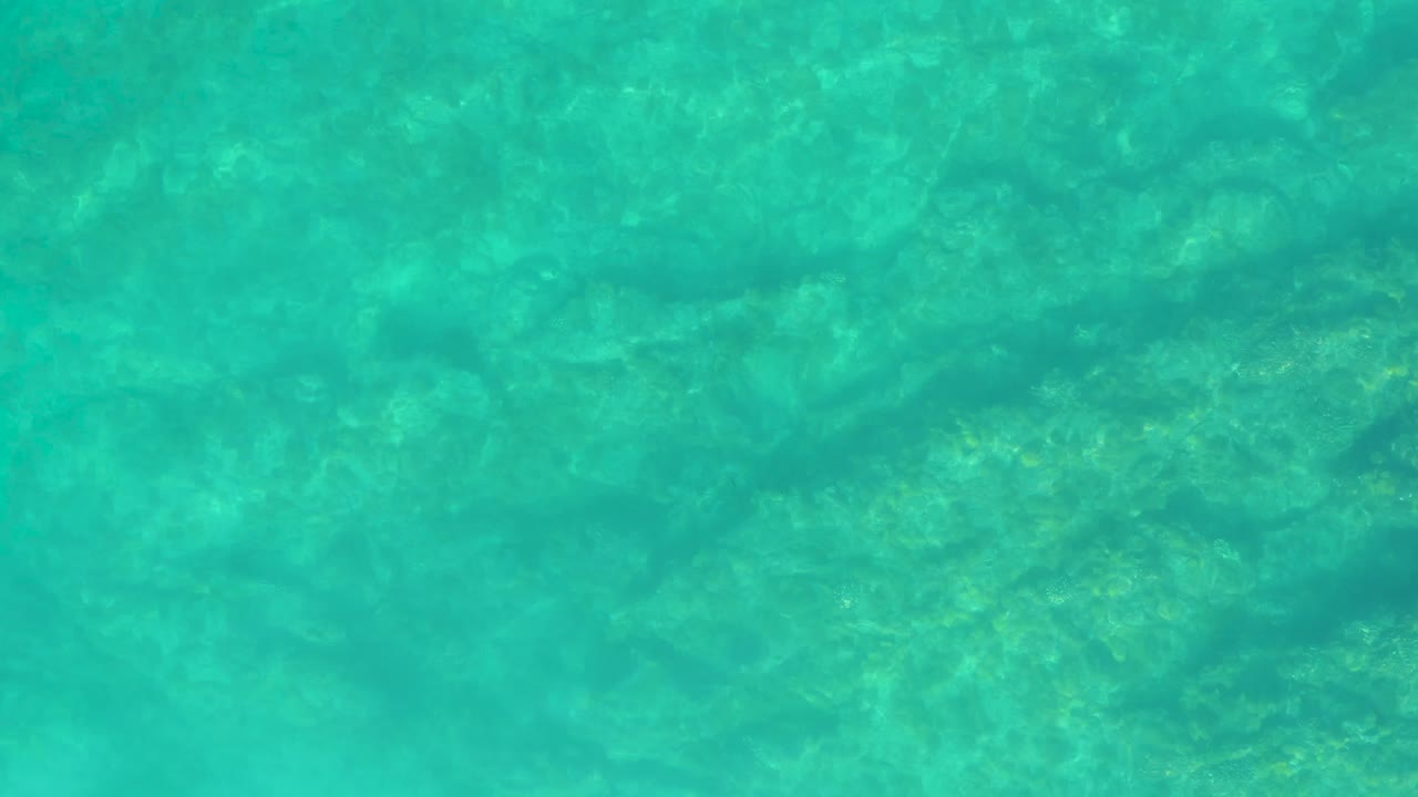 Clear turquoise water at Shipwrecks Los Cabos reveals darker rock patches and faint coral-like texture below the surface, with no visible people.