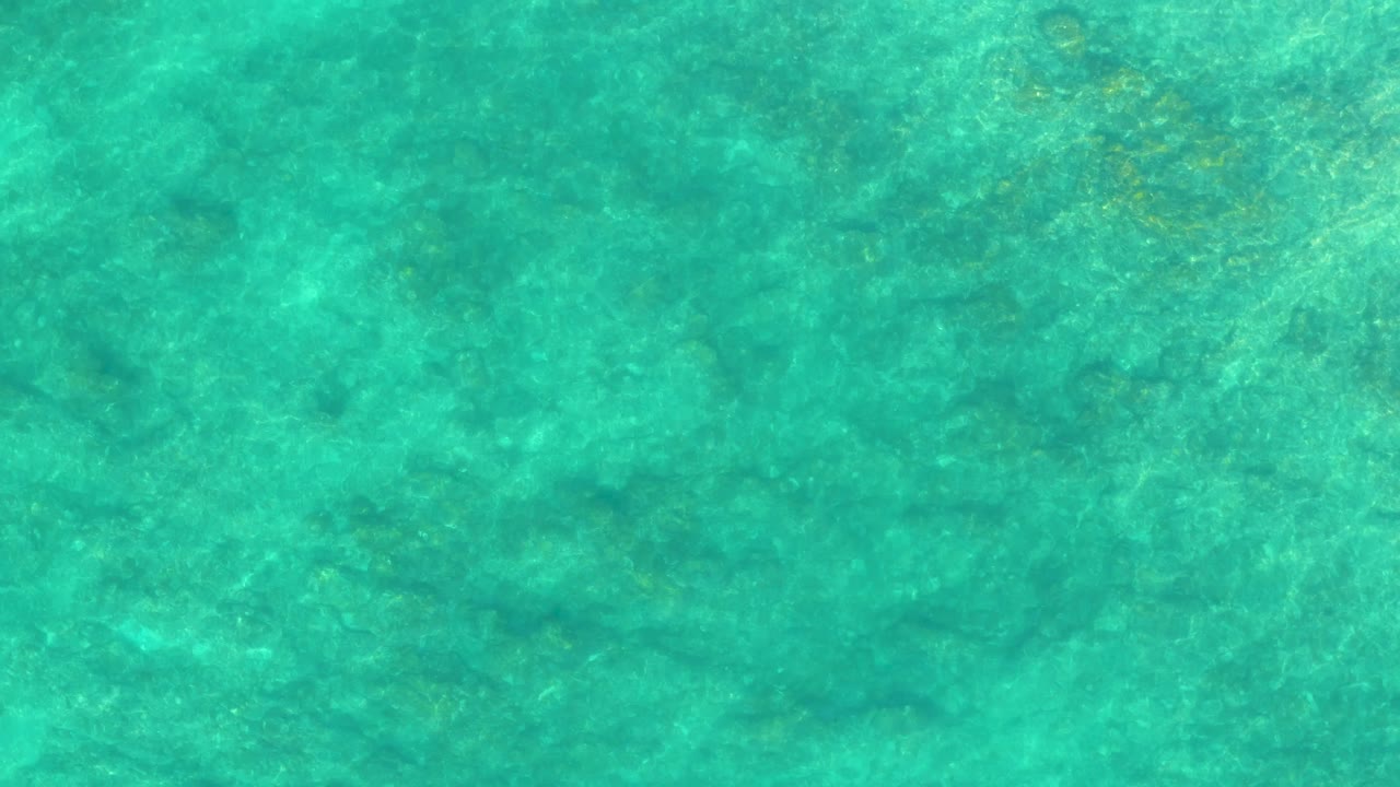 A full-frame overhead look at shallow turquoise seawater in Shipwrecks Los Cabos shows mottled seabed detail beneath calm surface motion and no visible people.