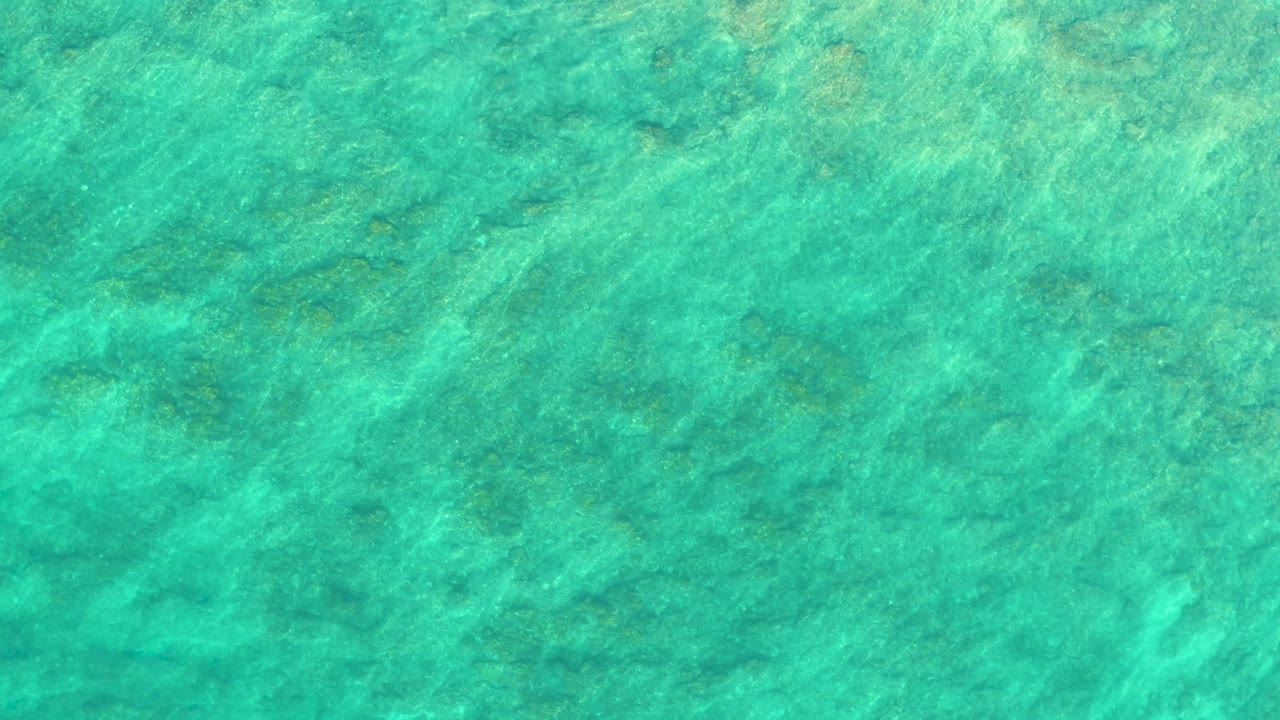 Second overhead frame of clear blue-green water at Shipwrecks Los Cabos, where faint wave streaks cross the visible seabed and no visible people appear.