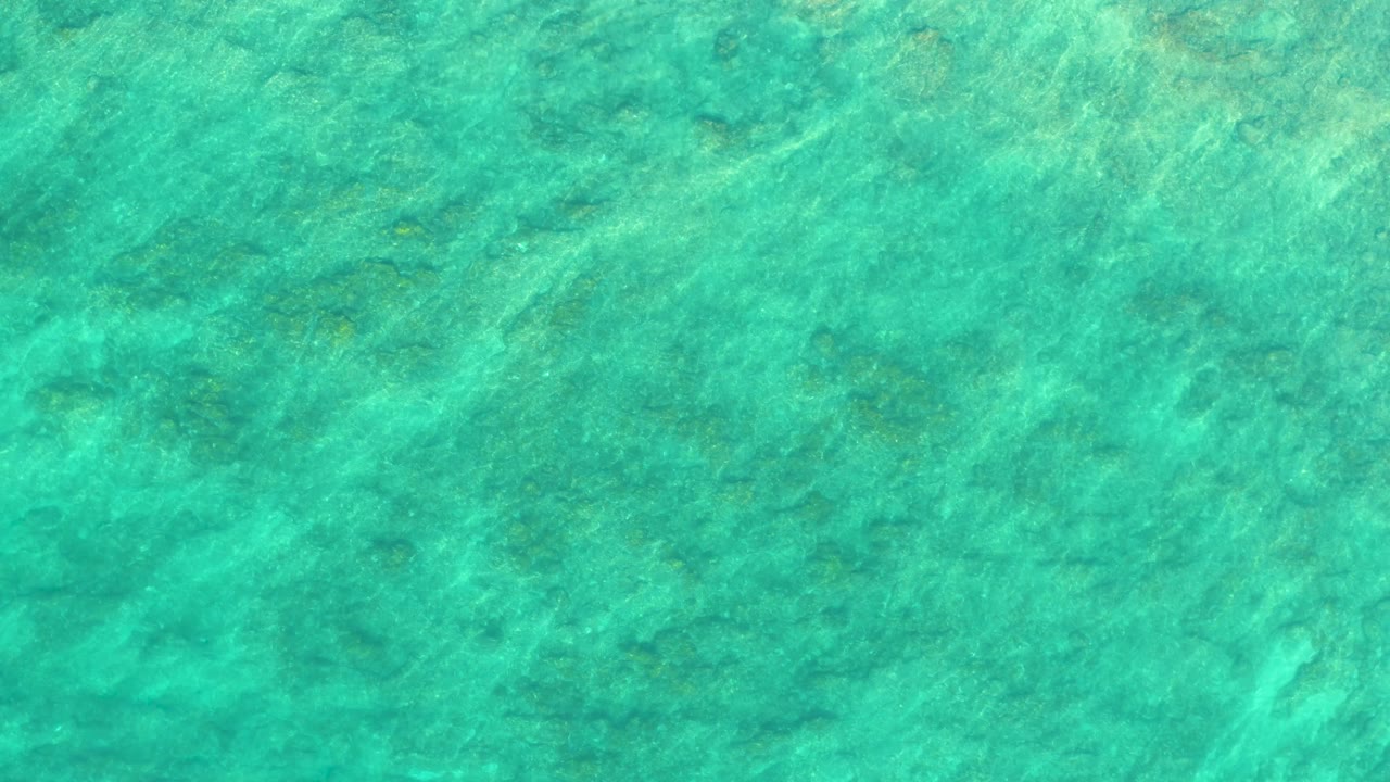 Overhead turquoise water frame from Shipwrecks Los Cabos, with pale sand or coral-like seabed patches softened by surface movement and no visible people.