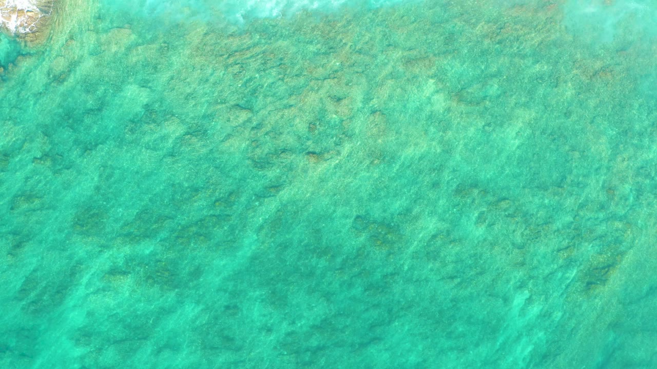Green-blue shallow water at Shipwrecks Los Cabos shows mottled seabed patterns and submerged coral under a slightly different ripple pattern, with no visible people.