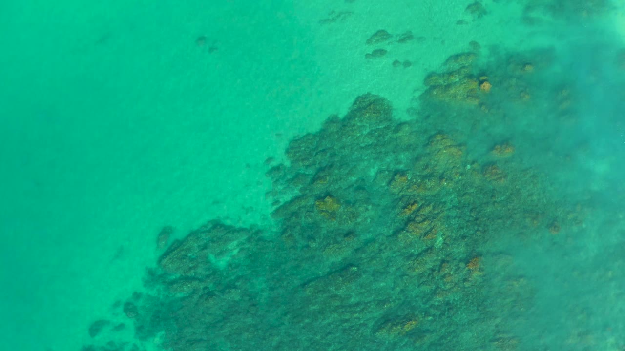 Turquoise water over a dark reef formation at Shipwrecks Los Cabos, with sandy seabed around the upper frame and no visible people.