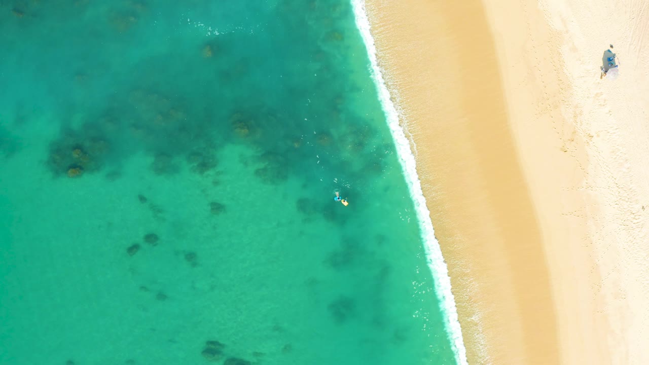 Top-down view at Shipwrecks Los Cabos showing turquoise shallow water, a narrow surf line, pale sand, dark underwater patches, and a small person on the upper beach.