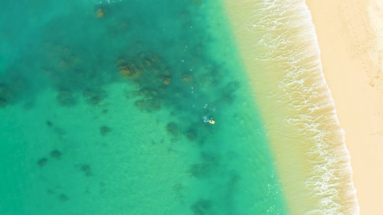 Overhead view at Shipwrecks Los Cabos showing turquoise water, darker reef patches, pale sand, a thin wave line, and a tiny swimmer near the shallows.
