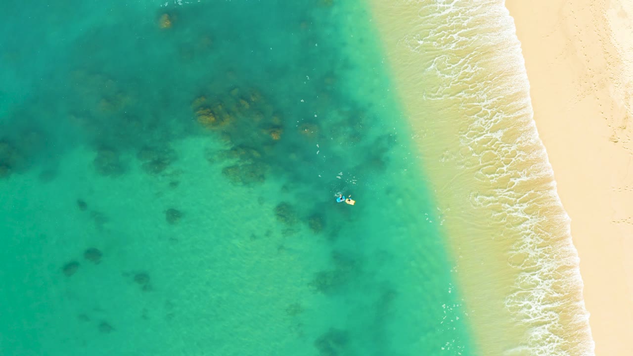At 0:01, the Shipwrecks Los Cabos shoreline remains framed from above, with the swimmer slightly lower in the water and the foam line edging the pale sand.